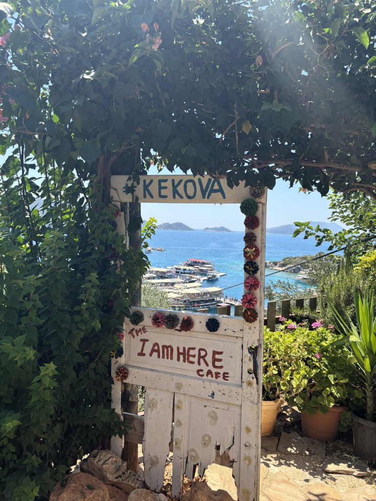 Kekova'ya açılan bir kapı, üzerinde 'Kekova' ve 'I AM HERE Cafe' yazılı, etrafında yeşil bitkilerle süslenmiş.